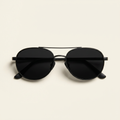 Manon oval aviator sunglasses in matte black with dark lenses, against a cream studio backdrop